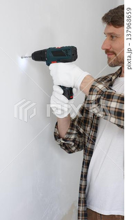 Middle aged male worker wearing checked shirt and white gloves, is drilling hole in a wall with cordless drill during home renovation work. Vertical portrait view Middle aged male worker wearing checked shirt and white gloves, is drilling hole in a wall with cordless drill during home renovation work. Vertical portrait view 137968659
