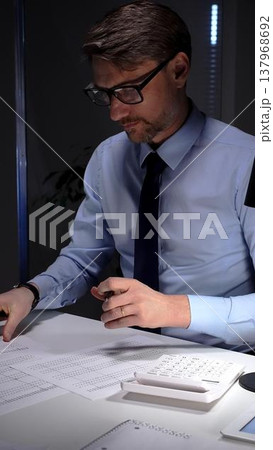 Businessman diligently auditing financial documents with a magnifying glass under focused lamp light. Audit and taxes in business Businessman diligently auditing financial documents with a magnifying glass under focused lamp light. Audit and taxes in business 137968692