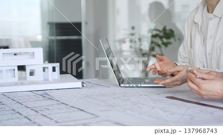 Architects collaborating on residential construction plans, reviewing blueprints and design models while working with laptop and architectural tools in a modern office 137968743