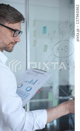 Young professional in glasses mapping business strategy and analyzing charts on a glass whiteboard, holding a clipboard while planning project goals, growth and execution Young professional in glasses mapping business strategy and analyzing charts on a glass whiteboard, holding a clipboard while planning project goals, growth and execution 137968862