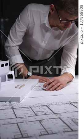 Architect examining blueprints and physical model, planning new construction project development. Concept of architecture and business in low key lighting Architect examining blueprints and physical model, planning new construction project development. Concept of architecture and business in low key lighting 137968908