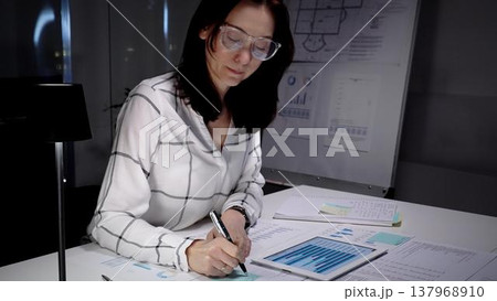Businesswoman working late night, analyzing data and financial reports using a digital tablet and taking notes in a dark modern office, focused on project deadline Businesswoman working late night, analyzing data and financial reports using a digital tablet and taking notes in a dark modern office, focused on project deadline 137968910
