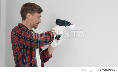 Middle aged male worker wearing red checked shirt and white gloves, is drilling hole in a wall with cordless drill during home renovation work. Portrait view Middle aged male worker wearing red checked shirt and white gloves, is drilling hole in a wall with cordless drill during home renovation work. Portrait view 137968953