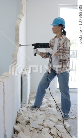 Female construction worker demolishing a wall with a hammer drill, wearing protective gear, generating rubble pile during renovation project 137968988