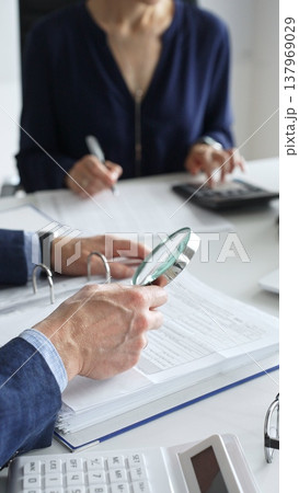 Business colleagues are working together, analyzing financial documents with a magnifying glass, calculating and taking notes in the office. Audit and taxes in business 137969029
