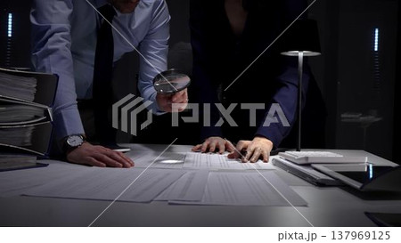 Business professionals collaborating over financial reports and a calculator beneath a desk lamp, reviewing documents and data late into the night to plan budget and strategy 137969125