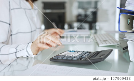 Woman calculating finances with black calculator, managing taxes, invoices, and accounting documents in office. Audit and taxes in business Woman calculating finances with black calculator, managing taxes, invoices, and accounting documents in office. Audit and taxes in business 137969438