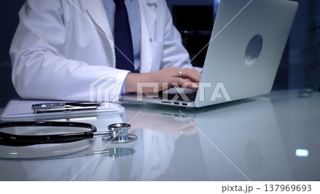 A stethoscope lies on a white reflective table while a male doctor works in a clinic office.. Wearing lab coat. Typing on laptop and writing notes. A stethoscope lies on a white reflective table while a male doctor works in a clinic office.. Wearing lab coat. Typing on laptop and writing notes. 137969693