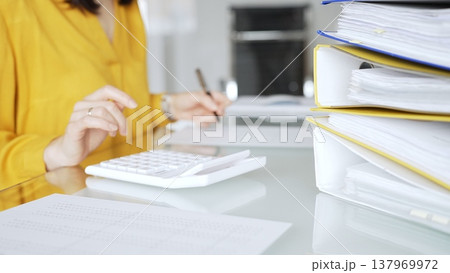 Woman in yellow blouse calculating finances at office desk, using calculator and writing, surrounded by documents and binders. Audit and taxes in business Woman in yellow blouse calculating finances at office desk, using calculator and writing, surrounded by documents and binders. Audit and taxes in business 137969972