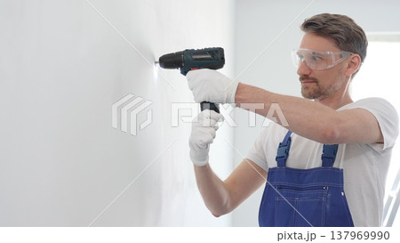 Professional handyman wearing blue overall, safety glasses and gloves, is using cordless drill to make hole in white wall during renovation or installation work 137969990