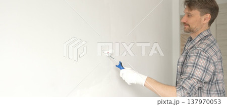 Man professional painter wearing checkered shirt, is rolling white paint on apartment wall, transforming interior space with steady strokes during comprehensive home renovation Man professional painter wearing checkered shirt, is rolling white paint on apartment wall, transforming interior space with steady strokes during comprehensive home renovation 137970053