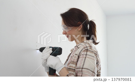 Woman wearing protective gear drilling hole in white wall during home improvement project, focused on precise home renovation work 137970081