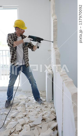 Man construction worker demolishing concrete wall with rotary hammer, wearing safety gear and yellow hardhat, generating rubble and dust during extensive home renovation process Man construction worker demolishing concrete wall with rotary hammer, wearing safety gear and yellow hardhat, generating rubble and dust during extensive home renovation process 137970092