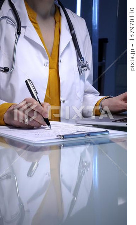Doctor writing patient medical records, managing data, and documenting information on a clipboard in a clinic setting, utilizing modern technology with a laptop Doctor writing patient medical records, managing data, and documenting information on a clipboard in a clinic setting, utilizing modern technology with a laptop 137970110