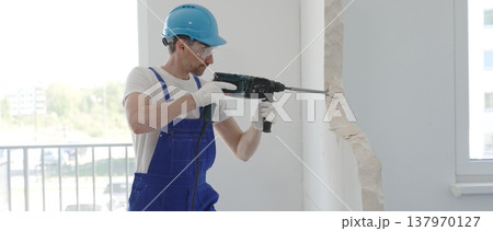 Male construction worker demolishing concrete wall with rotary hammer, wearing work overalls and blue hardhat is generating dust and debris during home renovation work, portrait horizontal view 137970127
