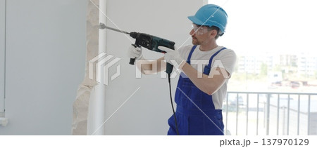 Construction worker demolishing concrete wall with rotary hammer, wearing protective safety gear, generating dust and debris during home renovation work, portrait horizontal view Construction worker demolishing concrete wall with rotary hammer, wearing protective safety gear, generating dust and debris during home renovation work, portrait horizontal view 137970129