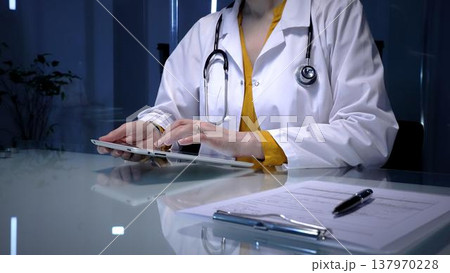 Female doctor wearing a white lab coat and stethoscope, using a digital tablet for telemedicine consultation, reviewing patient records, or managing hospital information system Female doctor wearing a white lab coat and stethoscope, using a digital tablet for telemedicine consultation, reviewing patient records, or managing hospital information system 137970228