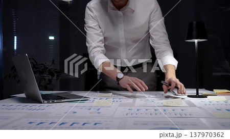 Professional businessman standing at a brightly lit desk, focused on writing notes and reviewing financial reports, charts and data late into the night to meet deadlines Professional businessman standing at a brightly lit desk, focused on writing notes and reviewing financial reports, charts and data late into the night to meet deadlines 137970362
