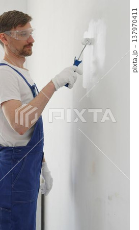 Man professional painter wearing protective white gloves and blue work overalls, applying paint with roller on pristine interior wall during home renovation project, vertical view Man professional painter wearing protective white gloves and blue work overalls, applying paint with roller on pristine interior wall during home renovation project, vertical view 137970411