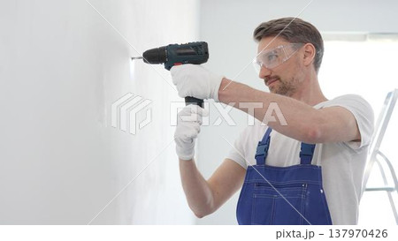 Middle aged man wearing blue overall and protective glasses, is drilling hole in white wall, wearing protective gear during home improvement project with cordless power tool 137970426