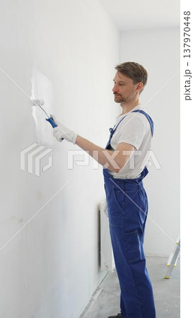 Male professional painter wearing protective white gloves and blue work overalls, applying paint with roller on pristine interior wall during home renovation project, vertical photo 137970448