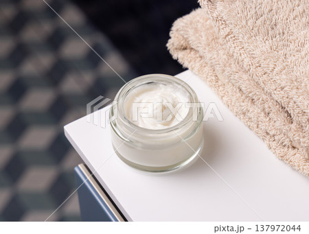 Opened cosmetic cream glass jar near beige bath towel against blue tiled floor top view Opened cosmetic cream glass jar near beige bath towel against blue tiled floor top view 137972044