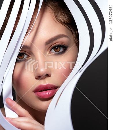 Art portrait of beautiful young woman with brown makeup. Beautiful brunette looks through trafaret paper. Model looking out through slots white paper. Art portrait of beautiful young woman with brown makeup. Beautiful brunette looks through trafaret paper. Model looking out through slots white paper. 137972484