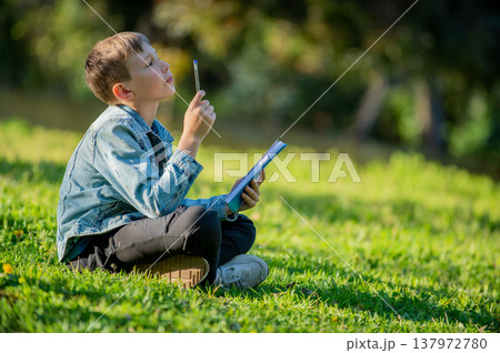 Thinking student with a textbook in his hands is sitting on a green lawn in a park. Pupil reading his notes and memorizing the text before a test. School boy writing in a notebook in a garden 137972780