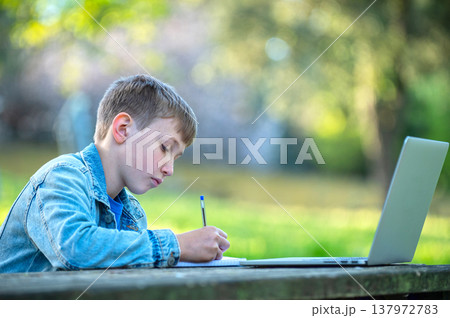 Boy is writing down notes outdoors. Scholar studying online with a computer. Boy on online lessons outside.  Student with a laptop and a notebook writing behind a table in a park. 137972783