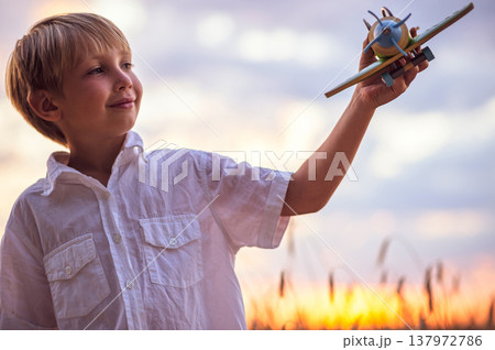 Happy child playing with a toy plane in nature during summer sunset. Boy in a  white shirt with a plane in hands on wheat field. Kid holds a wooden airplane and dreams of being a pilot, on the nature 137972786