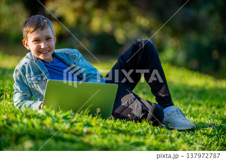 A happy Student kid with a laptop sits on a green lawn. A young smiling child laying down and typing in the park. Cheerful Young boy studying online with a computer, outdoors. Looking at the camera. A happy Student kid with a laptop sits on a green lawn. A young smiling child laying down and typing in the park. Cheerful Young boy studying online with a computer, outdoors. Looking at the camera. 137972787