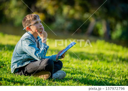 Thinking student with a textbook in his hands is sitting on a green lawn in a park. Young boy reading his notes and memorizing the text before a test. A student writing in a notebook in a garden Thinking student with a textbook in his hands is sitting on a green lawn in a park. Young boy reading his notes and memorizing the text before a test. A student writing in a notebook in a garden 137972796