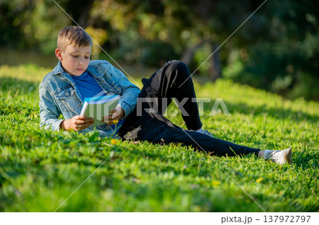 Schoolboy with a textbook in his hands is lying on a green lawn in a park during summer day. Studying teen looks to the notebook in a garden. Pupil reading his notes and memorizing the text outdoors 137972797