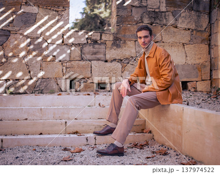 Spring fashion. Handsome male model in a brown leather jacket, beige trousers and sweater posing in an antique outdoor setting. Young man wearing stylish autumn clothes in brown and beige tones. 137974522