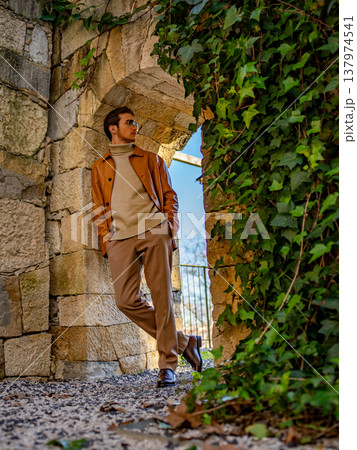 Attractive man wearing a brown leather jacket, beige trousers and a knit sweater posing in a vintage outdoor location. Male model posing in fashionable autumn outfit, brown and beige color palette. Attractive man wearing a brown leather jacket, beige trousers and a knit sweater posing in a vintage outdoor location. Male model posing in fashionable autumn outfit, brown and beige color palette. 137974541