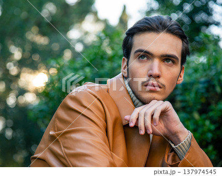Closeup portrait of  a stylish man wearing a elegance brown leather jacket and a knit sweater. Handsome male model posing in fashionable autumn, spring outfit in brown and beige tones, outdoors. 137974545