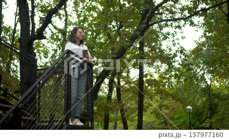 Young woman standing on a cafe veranda with a coffee cup, park backdrop. 137977160