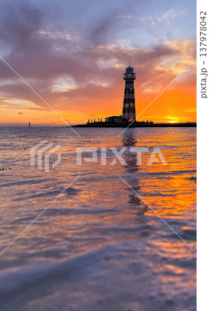 Vertical shot of a summer sunset. Sea foam in the foreground and the sun falling on the horizon with a lighthouse in the background. Vertical shot of a summer sunset. Sea foam in the foreground and the sun falling on the horizon with a lighthouse in the background. 137978042