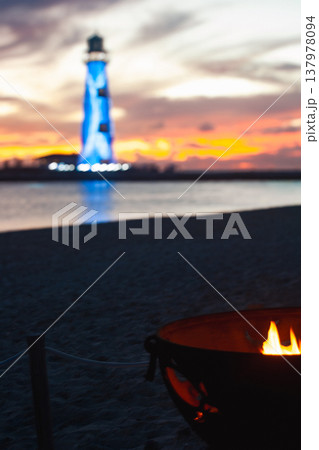 Beach bonfire casting warm light on sand, ocean, and a blue lighthouse reflecting dramatic sunset colors over the water, evoking a luxurious island getaway feeling 137978094