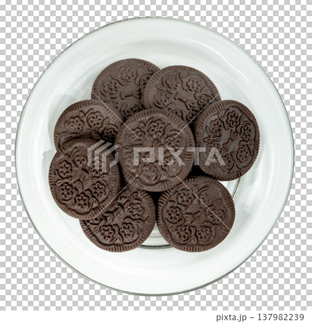 Top down view of a pile of dark chocolate sandwich cookies with floral patterns on a clear glass plate, isolated. Perfect for snack, dessert, or food industry marketing themes. 137982239