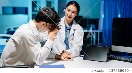 Asian doctor comforting stressed patient in hospital office, offering emotional support and professional mental health consultation with empathy and care. 137986080