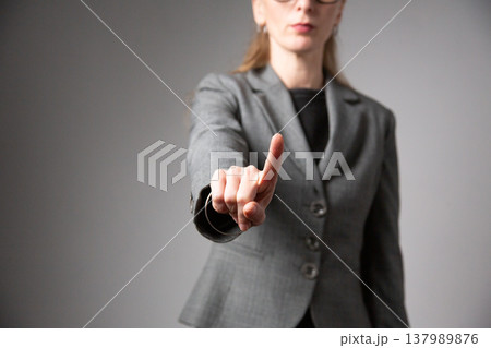 A portrait of a woman with glasses and long hair, wearing a business suit, extending her hand forward with her index finger raised. A prohibiting gesture. Vertical photo. A portrait of a woman with glasses and long hair, wearing a business suit, extending her hand forward with her index finger raised. A prohibiting gesture. Vertical photo. 137989876