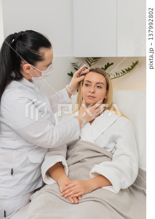 Cosmetologist in mask checking facial skin of young woman client before cosmetic treatment in clinic Cosmetologist in mask checking facial skin of young woman client before cosmetic treatment in clinic 137992802