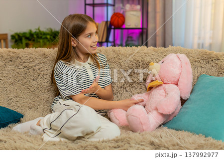 Child girl kid talking to plush bunny toy while sitting on home sofa in playful and imaginative mood Child girl kid talking to plush bunny toy while sitting on home sofa in playful and imaginative mood 137992977