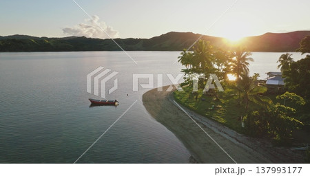 Small red boat anchored near a sandy beach with palm trees and houses, capturing the warm glow of the sun setting over calm ocean waters and distant hills in Fiji Small red boat anchored near a sandy beach with palm trees and houses, capturing the warm glow of the sun setting over calm ocean waters and distant hills in Fiji 137993177