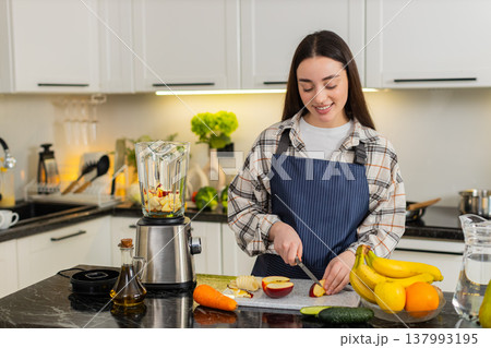 Young woman cuts apple for smoothie, preparing breakfast ingredients beside blender in home kitchen. 137993195