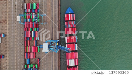 Cargo ship docks at a busy industrial port, having colorful shipping containers loaded and unloaded by cranes, representing global logistics and transportation. Top down aerial drone flight 137993369