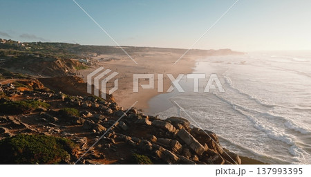 Portugal, Lisbon: Aerial view Praia do Guincho beach coastline with ocean waves rolling yellow sand, people relax and enjoy warm golden hour light sunset. Drone flight over rocky cliffs, zoom out 137993395