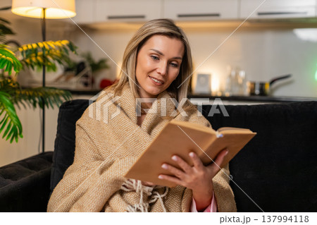 Adult woman wrapped in blanket reads favorite novel book relaxes enjoys quiet evening rest calm mood Adult woman wrapped in blanket reads favorite novel book relaxes enjoys quiet evening rest calm mood 137994118