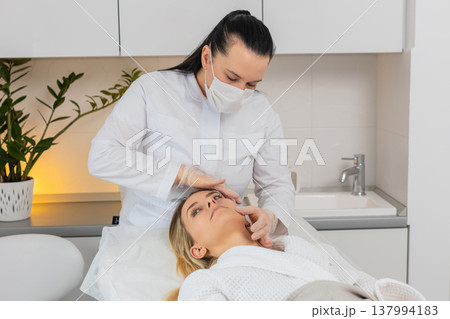 Young woman on couch while cosmetologist in gloves checks skin before relaxing cosmetic procedures Young woman on couch while cosmetologist in gloves checks skin before relaxing cosmetic procedures 137994183